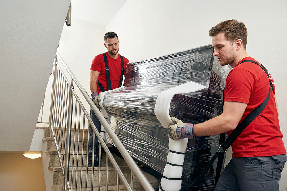 Dwuosobowa ekipa Boxperto Andrychów&nbsp;z&nbsp;pasami transportowymi wnosi zabezpieczone pianino po&nbsp;schodach w&nbsp;bloku bez windy.